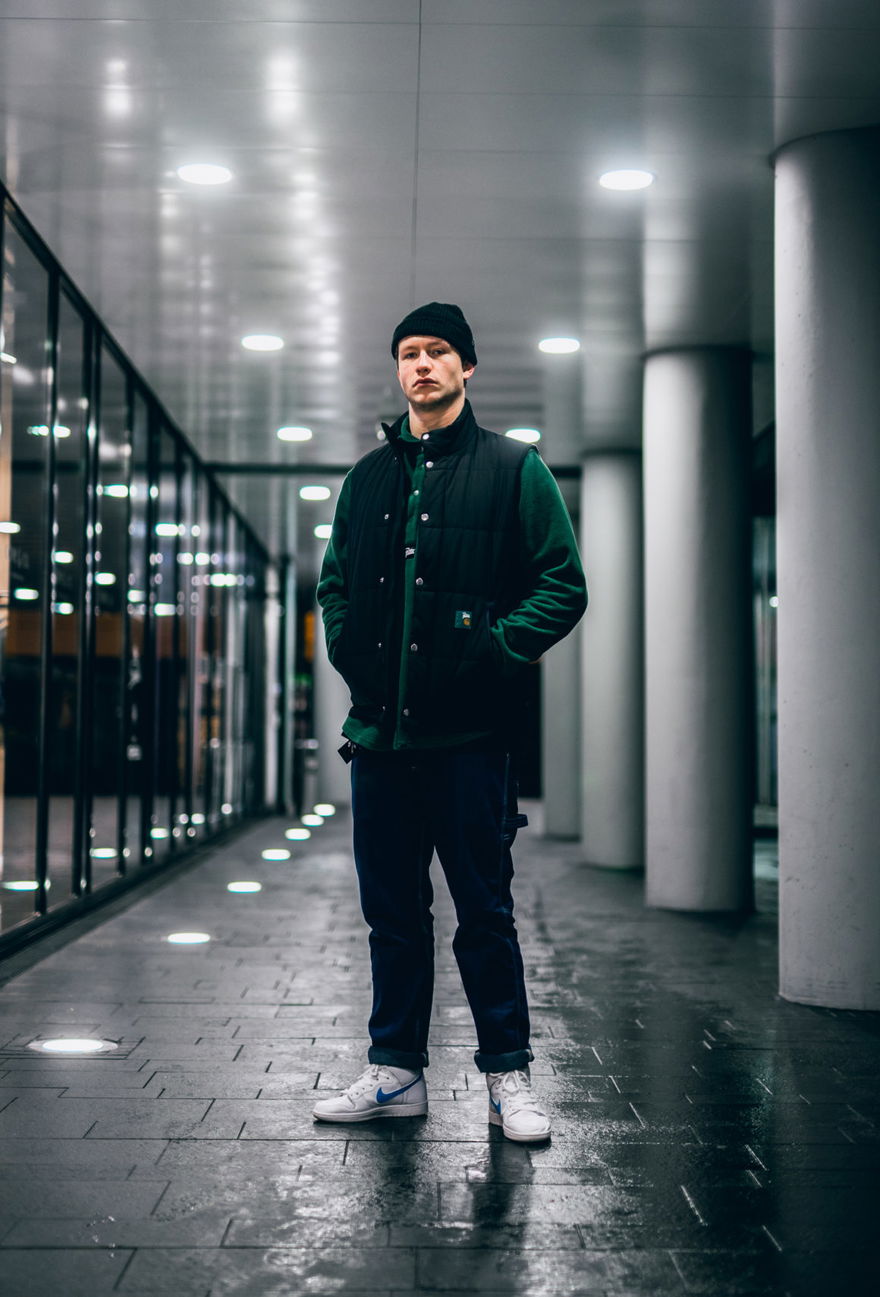 Porträt von themoldernway in einer nächtlichen Passage: Beanie, schwarze Steppweste, weiße Nike-Sneaker